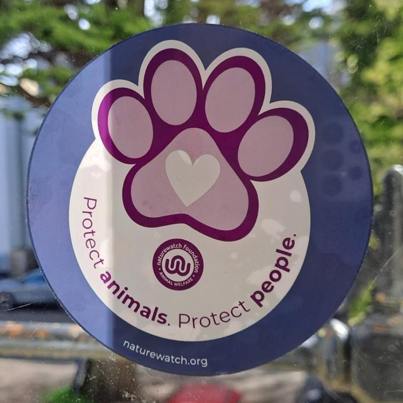Alt text: A circular Naturewatch Foundation sign attached to a post outdoors. The sign features a large purple paw print with a heart in the centre and the words “Protect animals. Protect people.” around the bottom.